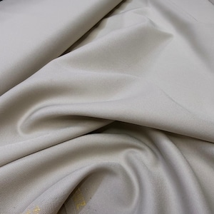 Puede incluir: Primer plano de una tela beige con pliegues suaves y textura sutil. El material parece ser un textil tejido, posiblemente para ropa o decoración del hogar. El color neutro y la caída sugieren una tela versátil.
