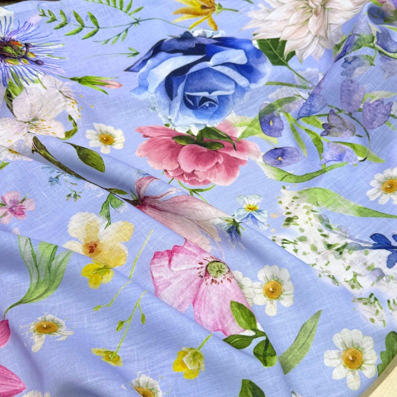 Peut inclure: Un tissu bleu clair avec un motif floral &agrave; l'aquarelle. Le motif pr&eacute;sente de grandes roses bleues, des pivoines roses, des marguerites blanches et d'autres fleurs color&eacute;es et des feuilles vertes. Le tissu est pli&eacute;, montrant la texture et les d&eacute;tails de l'impression.