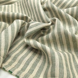 May include: Close-up of a folded fabric with a striped pattern. The fabric features alternating vertical stripes of off-white and dark green. The material appears to be woven, with a slightly textured surface and visible threads.
