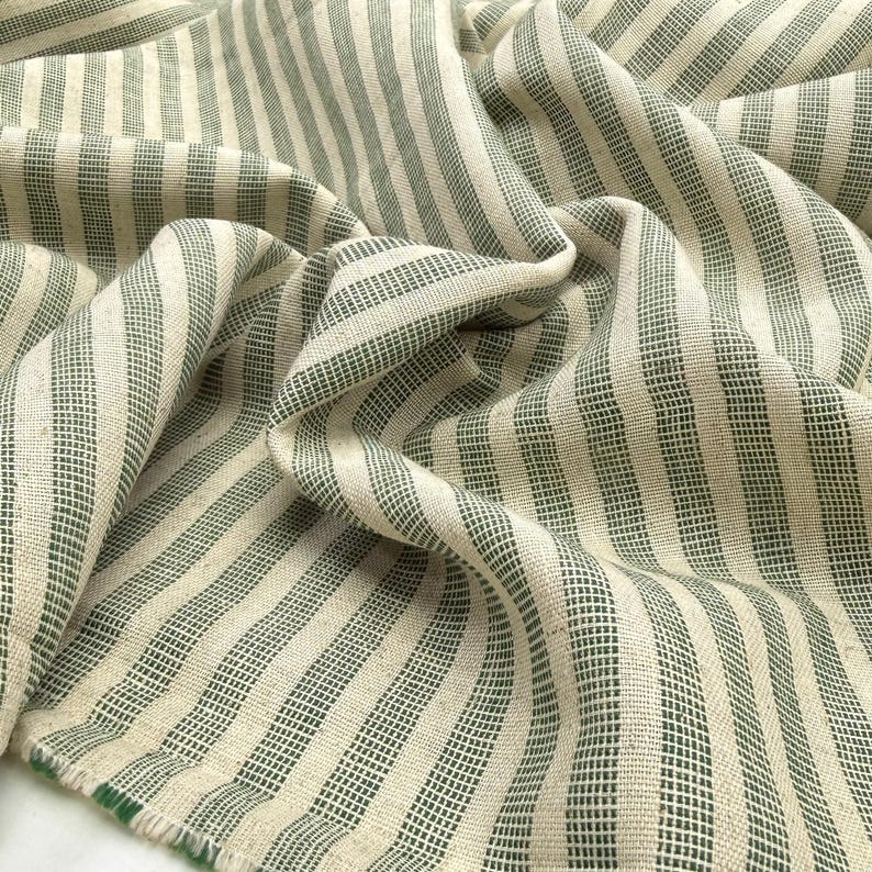 May include: Close-up of a folded fabric with a striped pattern. The fabric features alternating vertical stripes of off-white and dark green. The material appears to be woven, with a slightly textured surface and visible threads.