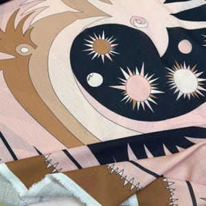 May include: A patterned fabric featuring a design with sun-like shapes, swirls, and geometric elements in shades of pink, brown, and navy blue. The fabric has a soft, flowing appearance, with a raw edge visible.