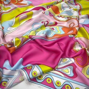 May include: A smooth fabric featuring a vibrant, multi-colored abstract pattern with bold swirls and shapes in shades of hot pink, lime green, bright orange, and light blue. The material has a soft, reflective sheen and a raw, unfinished edge.