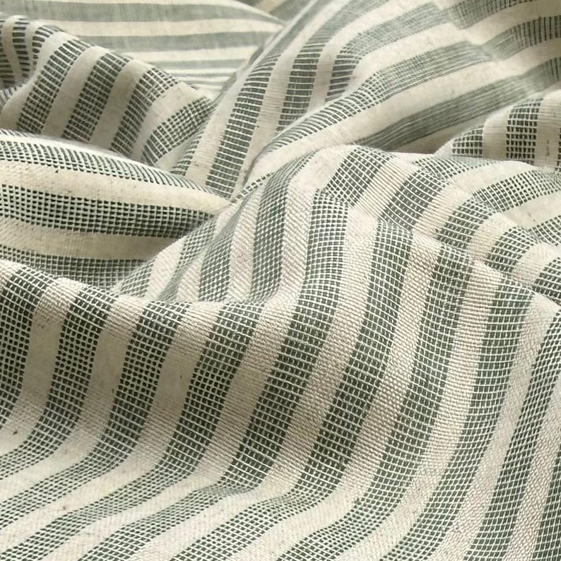 May include: Close-up of a textured fabric with a repeating pattern of vertical stripes. The stripes alternate between a light cream color and a muted green. The fabric is draped, creating soft folds and shadows.