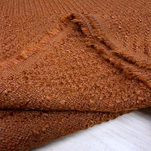 Könnte beinhalten: Nahaufnahme eines strukturierten Stoffes in einem warmen, gebrannten Orange. Der Stoff hat eine noppige Textur und einen Fransenrand, was auf eine Verwendung für Polster oder Kleidung hindeutet. Das Gewebe ist sichtbar und trägt zu seinem rustikalen Charme bei.