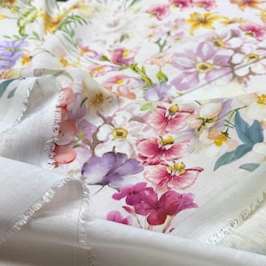 May include: White fabric with a vibrant floral pattern featuring pink, purple, yellow, and white flowers. The fabric has a delicate, soft appearance, with visible frayed edges, suggesting a textile or craft material.