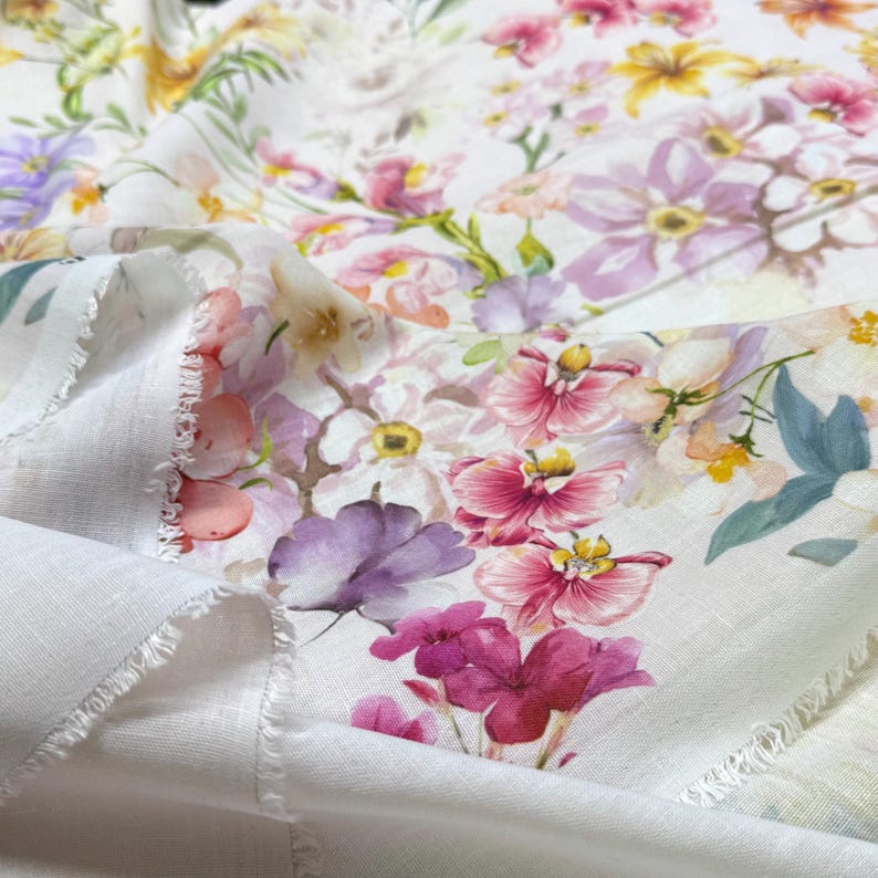 May include: White fabric with a vibrant floral pattern featuring pink, purple, yellow, and white flowers. The fabric has a delicate, soft appearance, with visible frayed edges, suggesting a textile or craft material.