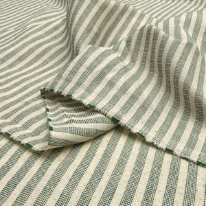 May include: Close-up of a folded fabric with a striped pattern. The fabric features alternating vertical stripes of off-white and green. The edges of the fabric are frayed, revealing the green color. The fabric appears to be linen or a similar natural fiber.