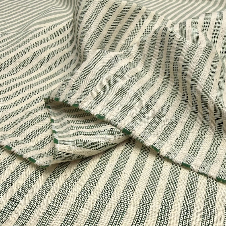 May include: Close-up of a folded fabric with a striped pattern. The fabric features alternating vertical stripes of off-white and green. The edges of the fabric are frayed, revealing the green color. The fabric appears to be linen or a similar natural fiber.