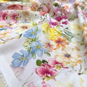 May include: Close-up of a white fabric with a vibrant floral pattern. The design features a variety of flowers in shades of pink, yellow, orange, and blue. The fabric is slightly wrinkled, showing the texture of the material.