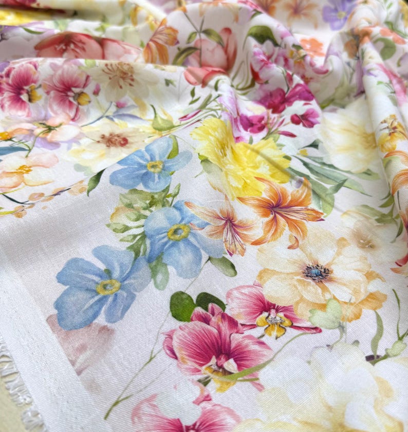 May include: Close-up of a white fabric with a vibrant floral pattern. The design features a variety of flowers in shades of pink, yellow, orange, and blue. The fabric is slightly wrinkled, showing the texture of the material.