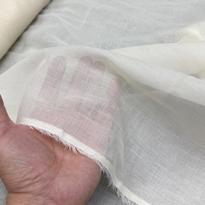 Könnte beinhalten: Ein transparenter, cremeweißer Stoff mit sichtbarer Webstruktur und einer ausgefransten, rohen Kante. Das Material ist leicht und durchscheinend, sodass der Hautton unter den feinen, halbtransparenten Textilfasern sichtbar ist.