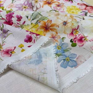 May include: White fabric with a vibrant floral pattern featuring pink, yellow, orange, and blue flowers. The fabric has a raw, unfinished edge, and the flowers are painted in a watercolor style. The fabric is folded, revealing the underside.