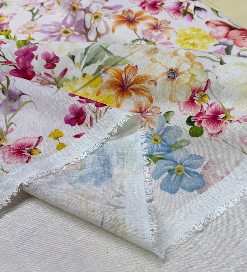 May include: White fabric with a vibrant floral pattern featuring pink, yellow, orange, and blue flowers. The fabric has a raw, unfinished edge, and the flowers are painted in a watercolor style. The fabric is folded, revealing the underside.