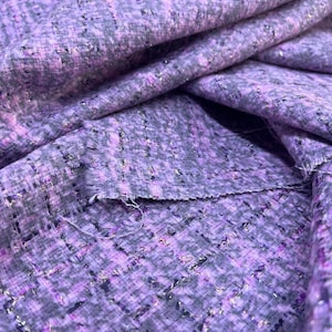Könnte beinhalten: Nahaufnahme eines strukturierten Stoffes in Lila-, Grau- und Silbertönen. Das gewebte Material hat ein tweedartiges Aussehen mit einer Mischung aus Farben und metallischen Fäden. Der Stoff ist gefaltet und zeigt die Textur und die Schichten.