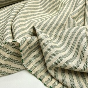 May include: Close-up of a folded fabric with a cream-colored background and green stripes. The fabric has a textured appearance and a raw edge with green trim. The stripes run diagonally across the fabric.