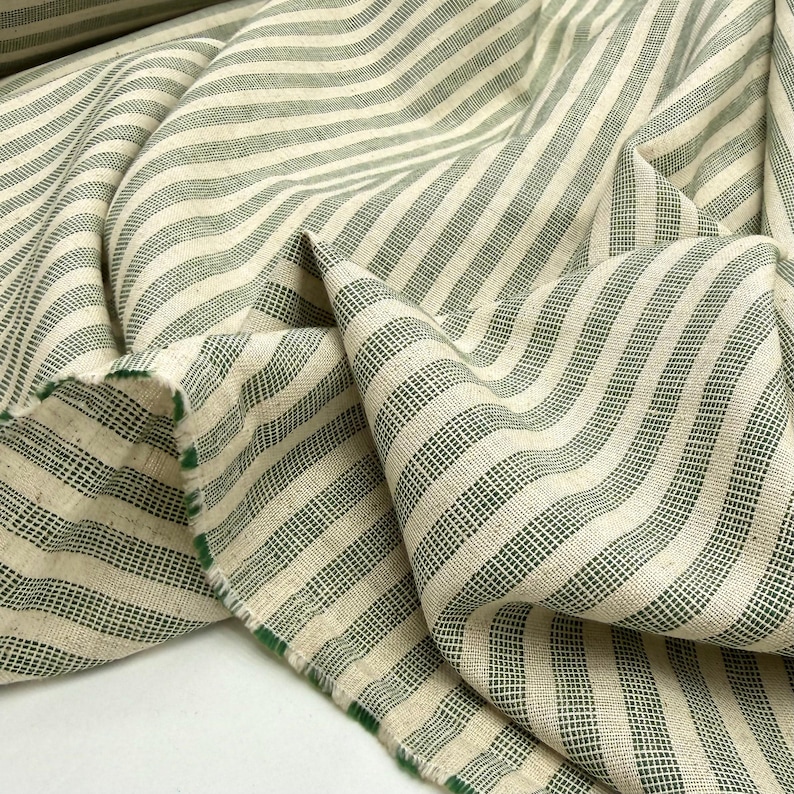 May include: Close-up of a folded fabric with a cream-colored background and green stripes. The fabric has a textured appearance and a raw edge with green trim. The stripes run diagonally across the fabric.