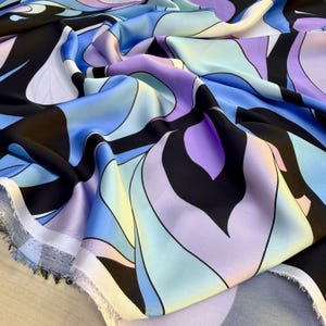 Op de afbeelding: Een stuk stof met een opvallend, abstract patroon van vloeiende, organische vormen in tinten blauw, lavendel, lichtgeel en zwart. Het materiaal heeft een gladde, satijnachtige afwerking met zichtbare plooien en een rafelige rand aan de onderkant.