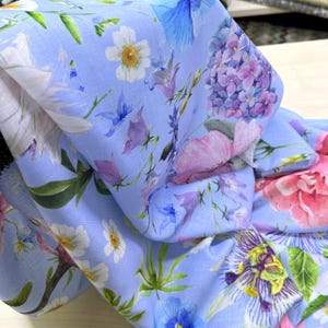 Peut inclure: Gros plan sur un tissu bleu clair orn&eacute; d'un motif floral. Le motif pr&eacute;sente diverses fleurs roses, violettes, blanches et bleues, avec des feuilles vertes. Le tissu est pli&eacute;, r&eacute;v&eacute;lant le motif floral r&eacute;p&eacute;t&eacute;.