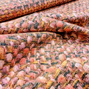 Puede incluir: Primer plano de una tela texturizada con un patrón tejido. La tela presenta una mezcla de hilos rosas, melocotón, amarillos y grises oscuros, creando un efecto complejo y multitonal. El material parece ser un tweed o un textil tejido similar.