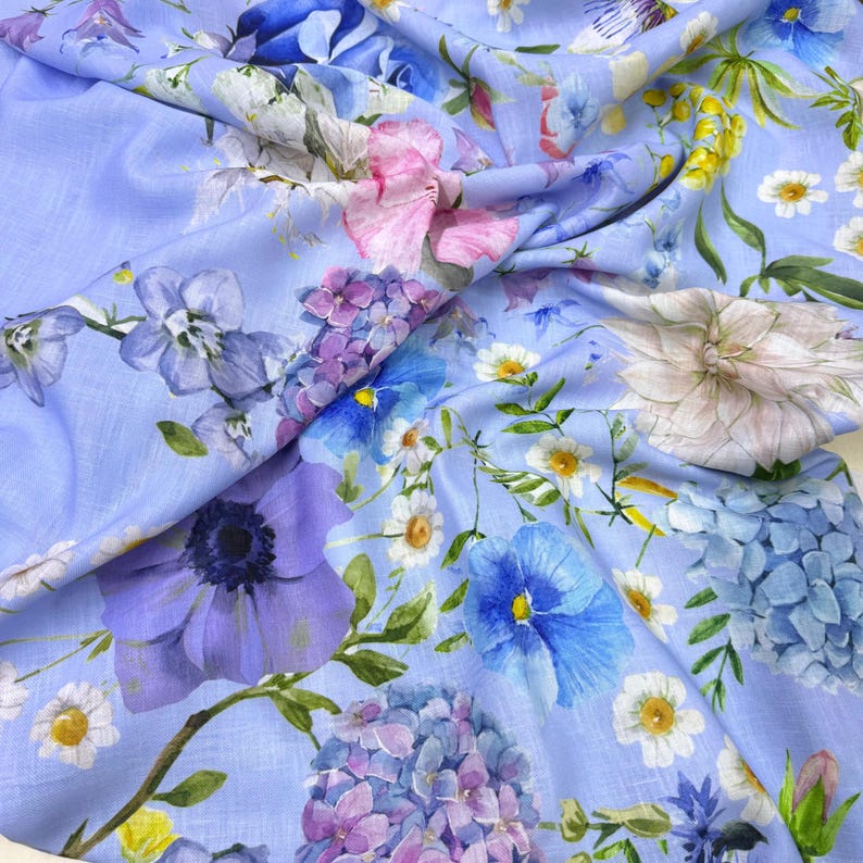 Peut inclure: Un tissu bleu clair orn&eacute; d'un motif floral. Le design pr&eacute;sente une vari&eacute;t&eacute; de fleurs dans des tons de bleu, violet, rose et blanc, notamment des hortensias, des an&eacute;mones et des marguerites. Des feuilles et des tiges vertes pars&egrave;ment le motif.