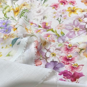 May include: White fabric with a vibrant floral pattern. The design features a variety of flowers in shades of pink, purple, yellow, and orange, set against a white background. The fabric has a soft, textured appearance.