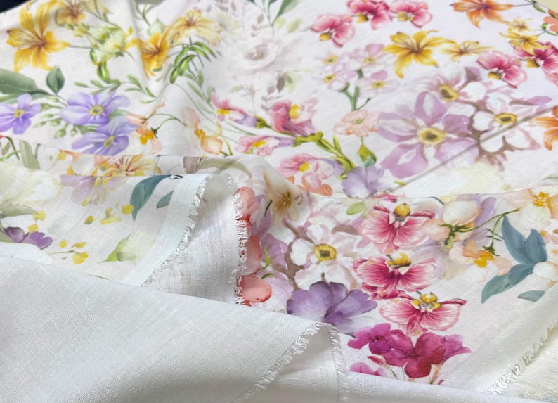 May include: White fabric with a vibrant floral pattern. The design features a variety of flowers in shades of pink, purple, yellow, and orange, set against a white background. The fabric has a soft, textured appearance.