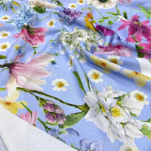 Peut inclure: Un tissu bleu clair orn&eacute; d'un motif floral. Le motif pr&eacute;sente une vari&eacute;t&eacute; de fleurs dans des tons de rose, de violet, de jaune et de blanc. Le tissu a un aspect doux et fluide, avec une bordure blanche visible sur le bord. Le motif comprend des illustrations botaniques d&eacute;taill&eacute;es.