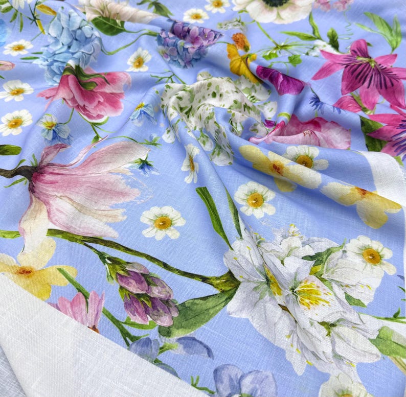 Peut inclure: Un tissu bleu clair orn&eacute; d'un motif floral. Le motif pr&eacute;sente une vari&eacute;t&eacute; de fleurs dans des tons de rose, de violet, de jaune et de blanc. Le tissu a un aspect doux et fluide, avec une bordure blanche visible sur le bord. Le motif comprend des illustrations botaniques d&eacute;taill&eacute;es.