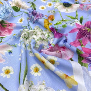 Peut inclure: Un tissu bleu clair orn&eacute; d'un motif floral color&eacute;. Le motif pr&eacute;sente diverses fleurs roses, violettes, jaunes et blanches. Le tissu semble doux et fluide, avec des plis et des plis visibles.