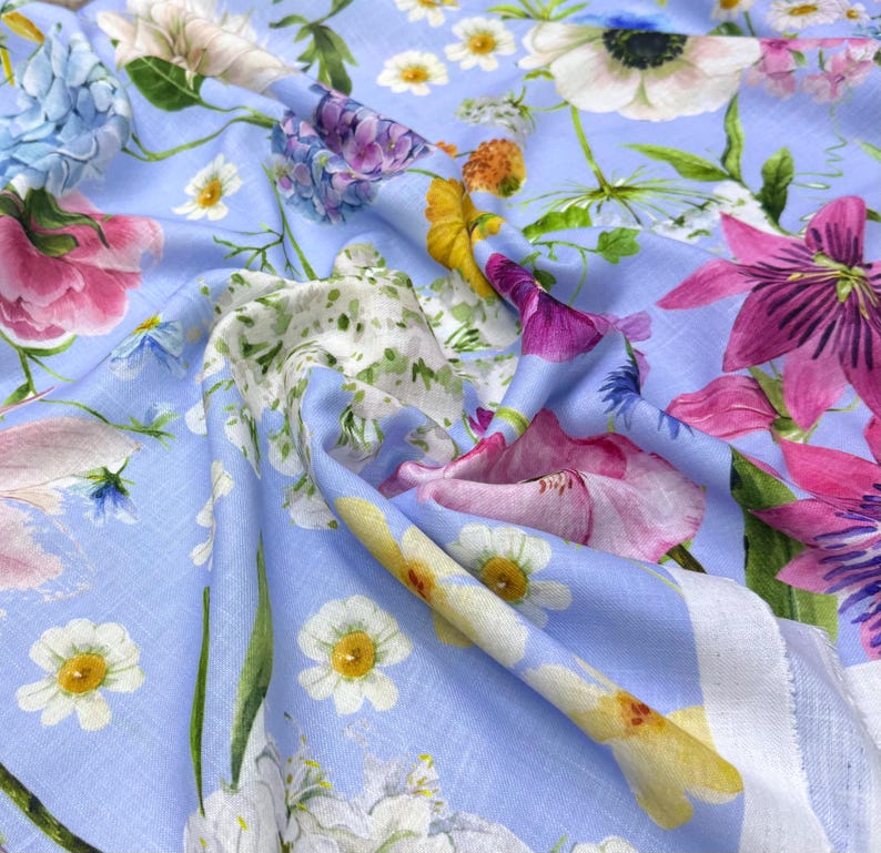 Peut inclure: Un tissu bleu clair orn&eacute; d'un motif floral color&eacute;. Le motif pr&eacute;sente diverses fleurs roses, violettes, jaunes et blanches. Le tissu semble doux et fluide, avec des plis et des plis visibles.
