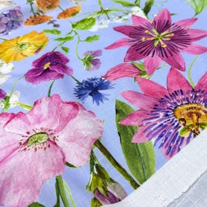 Peut inclure: Gros plan sur un tissu floral avec un fond bleu clair. Le tissu pr&eacute;sente une vari&eacute;t&eacute; de fleurs color&eacute;es, notamment des coquelicots roses, des passiflores violettes, des fleurs jaunes et des bleuets bleus. Des feuilles et des tiges vertes sont &eacute;galement visibles.