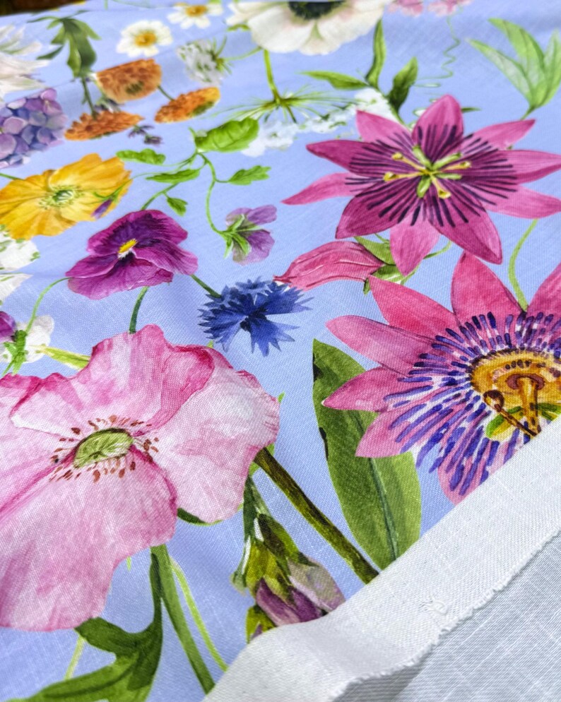 Peut inclure: Gros plan sur un tissu floral avec un fond bleu clair. Le tissu pr&eacute;sente une vari&eacute;t&eacute; de fleurs color&eacute;es, notamment des coquelicots roses, des passiflores violettes, des fleurs jaunes et des bleuets bleus. Des feuilles et des tiges vertes sont &eacute;galement visibles.