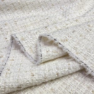 May include: A white tweed fabric featuring a textured weave with interwoven metallic gold threads. The material displays a frayed, raw edge along the folded section, showcasing a loose, multi-layered construction with a visible black thread.