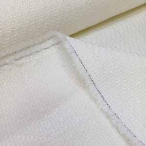 May include: Close-up of a textured, off-white fabric. The material has a slightly raised, nubby surface and a soft, fringed edge. The fabric is folded, revealing a glimpse of the rolled-up portion in the background. The overall appearance is clean and elegant.