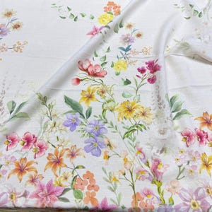 May include: White fabric with a vibrant floral pattern. The design features a variety of flowers in shades of pink, yellow, orange, and purple, with green leaves and stems. The fabric is folded, showing the detailed print.