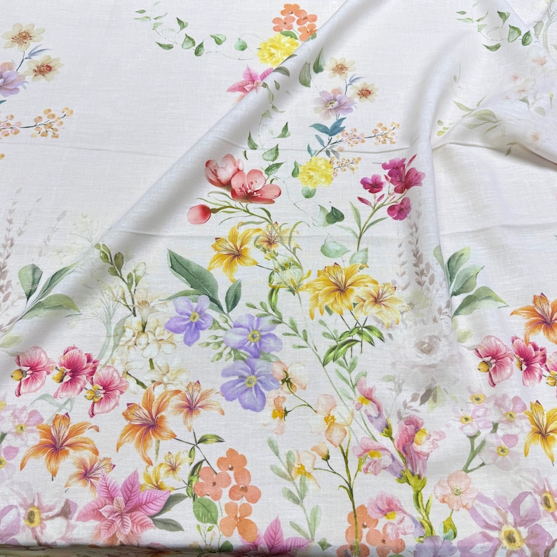 May include: White fabric with a vibrant floral pattern. The design features a variety of flowers in shades of pink, yellow, orange, and purple, with green leaves and stems. The fabric is folded, showing the detailed print.