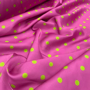 May include: A vibrant hot pink fabric featuring a repeating pattern of small, evenly spaced lime green polka dots. The material has a smooth, slightly reflective surface with soft folds and drapes that create natural shadows across the textile.