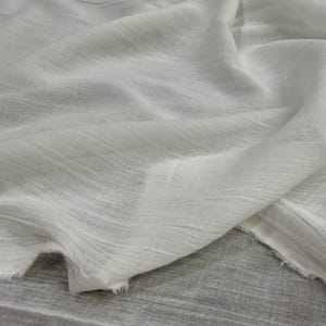以下が含まれることがあります： 目に見えるスラブテクスチャーと半透明の品質を持つ白い生地。素材は柔らかいひだにドレープされており、テキスタイルの底辺と側面に沿って未加工の切りっぱなしの端が見える軽量な外観を特徴としています。