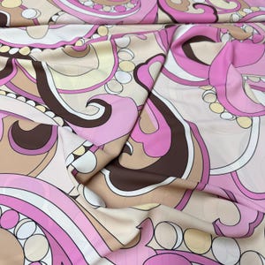 May include: A close-up of a fabric with a retro-style pattern. The design features swirling shapes in shades of pink, brown, white, and beige. The fabric is folded, showing the pattern's repeating nature and the fabric's texture.