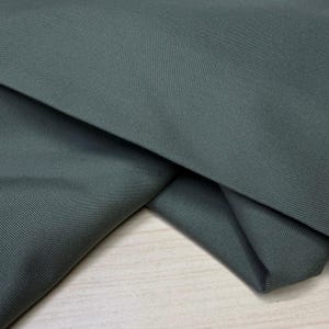 Puede incluir: Primer plano de una tela doblada en un tono verde azulado oscuro y apagado. La tela tiene una sutil textura acanalada, con líneas diagonales que recorren la superficie. Los pliegues crean sombras y profundidad, resaltando la textura del material.
