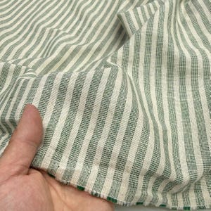May include: Close-up of a fabric with vertical stripes in white and green. The fabric is textured and appears to be cotton or linen. The stripes are evenly spaced and the fabric is slightly wrinkled.