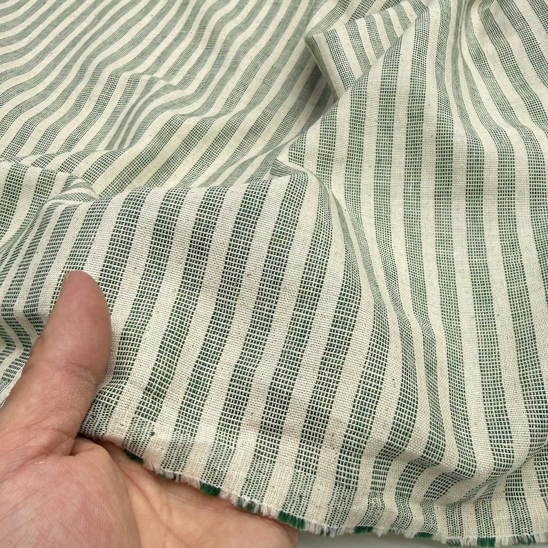 May include: Close-up of a fabric with vertical stripes in white and green. The fabric is textured and appears to be cotton or linen. The stripes are evenly spaced and the fabric is slightly wrinkled.