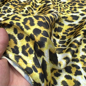 Puede incluir: Una pieza de tela con un vibrante estampado de leopardo en amarillo y negro. El material tiene una superficie lisa y ligeramente reflectante con pliegues que crean profundidad y textura en todo el diseño de manchas animales.