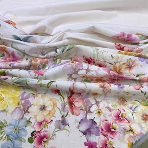 May include: White fabric with a vibrant floral pattern featuring a variety of flowers in shades of pink, yellow, orange, purple, and blue. The fabric is draped, showing folds and texture, with a plain white fabric in the background.