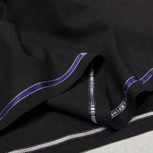 May include: A close-up shot of black fabric, folded to show texture and depth. The edge features a blue and white stripe with text. The material suggests a high-quality textile, suitable for clothing.