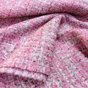 May include: Close-up of pink, white, and gray tweed fabric. The fabric has a textured weave with flecks of metallic thread, creating a shimmering effect. The fabric is folded, showing the texture and color variations.