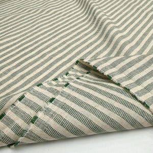 May include: Close-up of a folded fabric with a striped pattern. The fabric features alternating stripes of off-white and dark green. The edges of the fabric are frayed, revealing the texture of the material. The fabric appears to be cotton or linen.