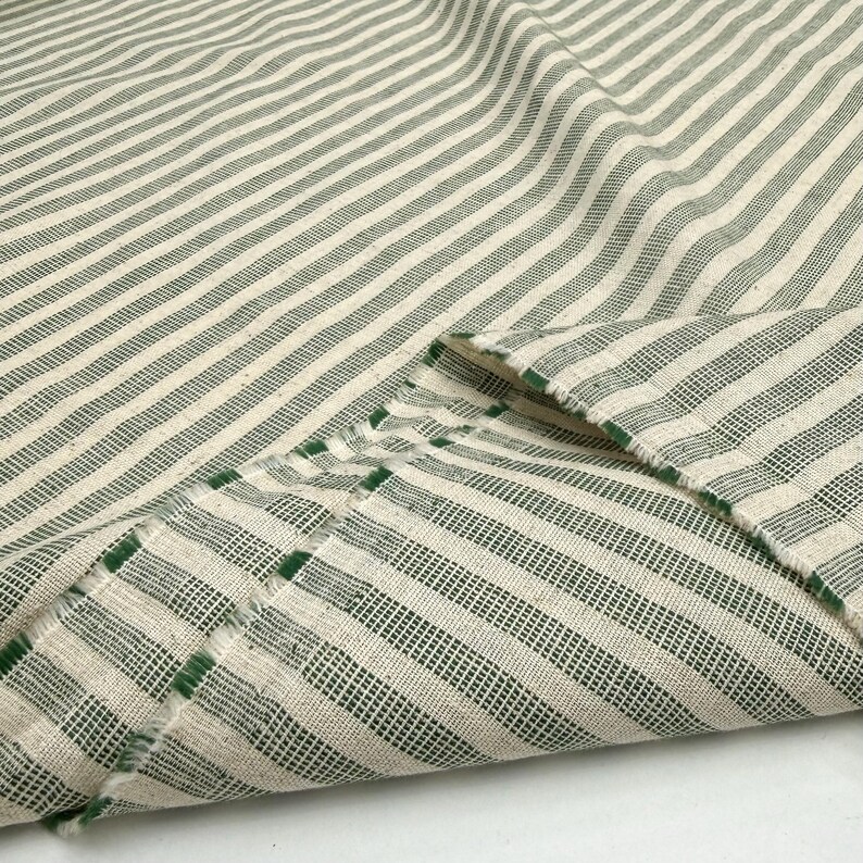 May include: Close-up of a folded fabric with a striped pattern. The fabric features alternating stripes of off-white and dark green. The edges of the fabric are frayed, revealing the texture of the material. The fabric appears to be cotton or linen.