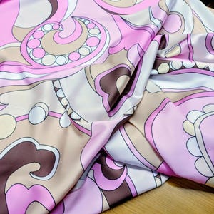 Puede incluir: Primer plano de una tela sedosa con un diseño retro. El patrón presenta formas arremolinadas en tonos de rosa, marrón y beige, con acentos blancos. La tela tiene un brillo brillante, lo que sugiere un material lujoso.