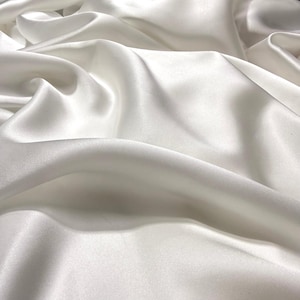 Puede incluir: Primer plano de una tela de satén blanco con pliegues suaves y un brillo lustroso. La tela parece suave y fluida, con sutiles variaciones de tono y textura. El efecto general es de lujo y elegancia.
